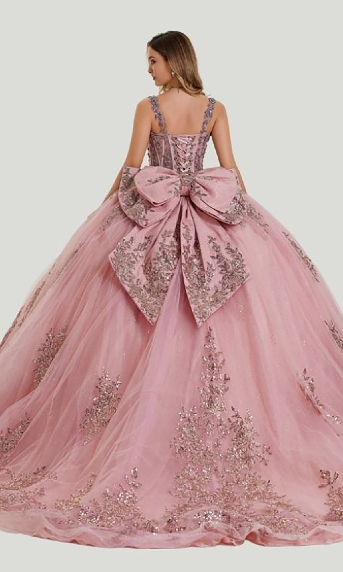A woman wearing the Quinceanera Dress 26406 by Stella Couture—a pink ball gown adorned with silver embroidery, a large bow at the back, delicate embroidered mesh, and glitter tulle—stands against a simple light background.